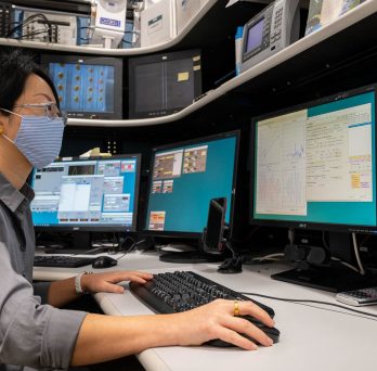 man wearing face mask looking at several computer monitors 